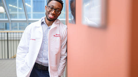Black, male medical student leaning against a wall while wearing a white coat